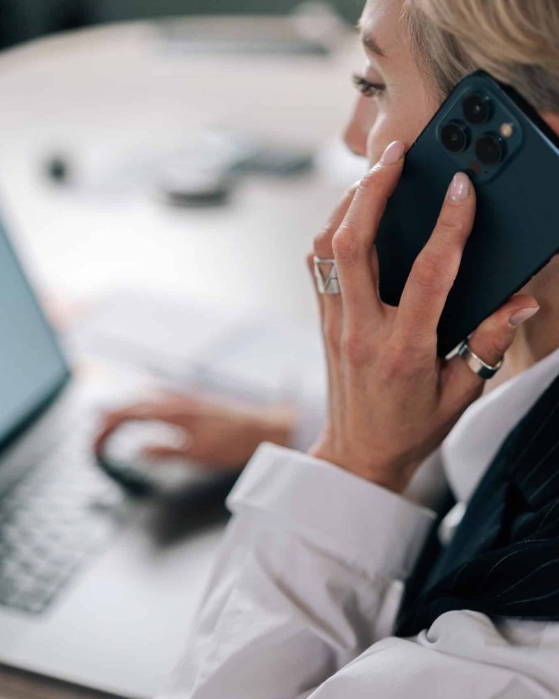 Closeup view from shoulder of confident senior adult 50s business woman talking on mobile phone at workplace table, sitting at laptop computer, discussing work, making call from workplace.
